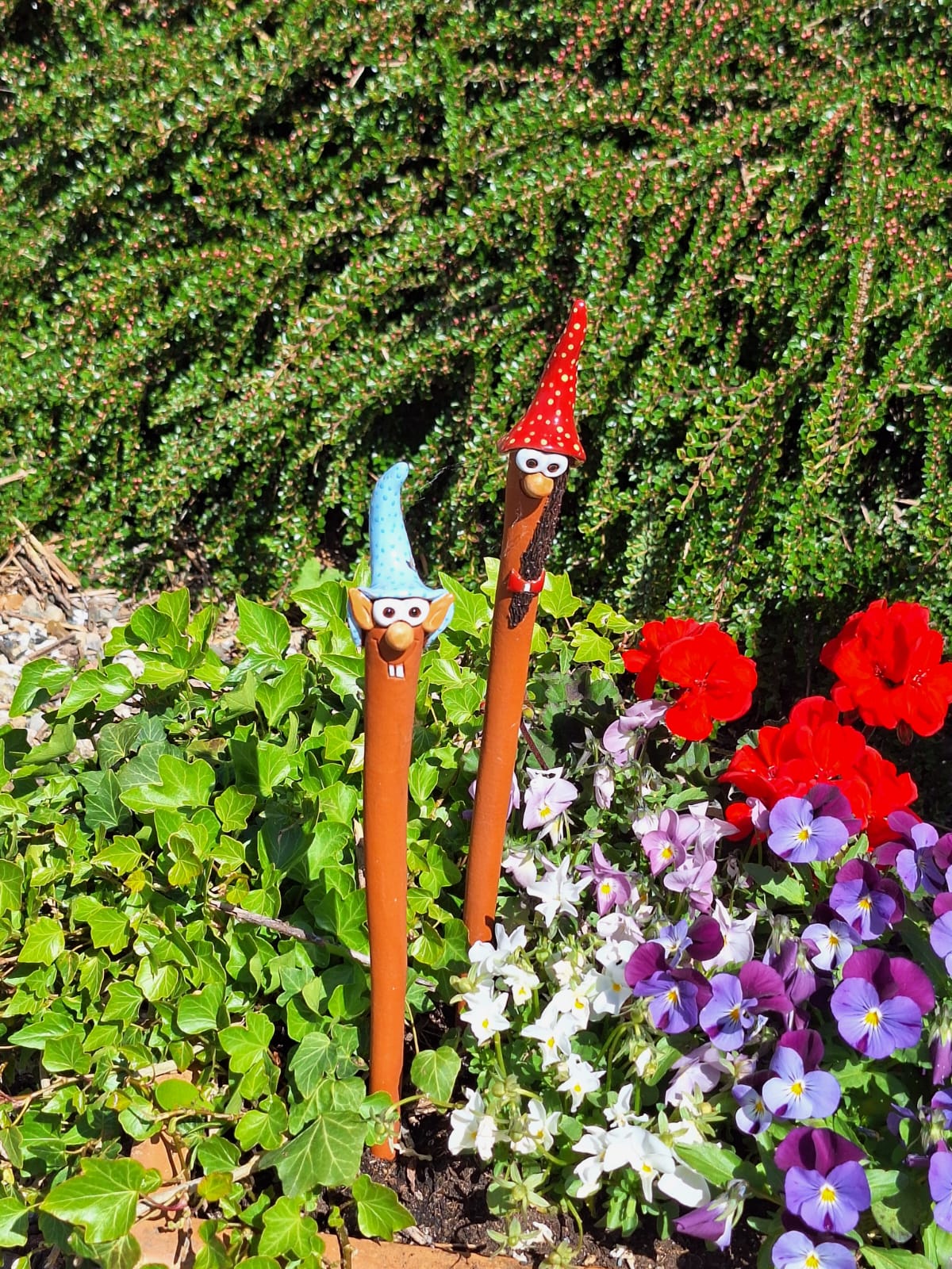 Tonfiguren mit langen Hälsen und bunten Zipfelmützen in Blumenbeet